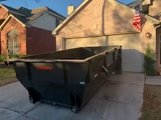Empty dark gray roll-off dumpster parked on the driveway of a brick house in front of the garage door.