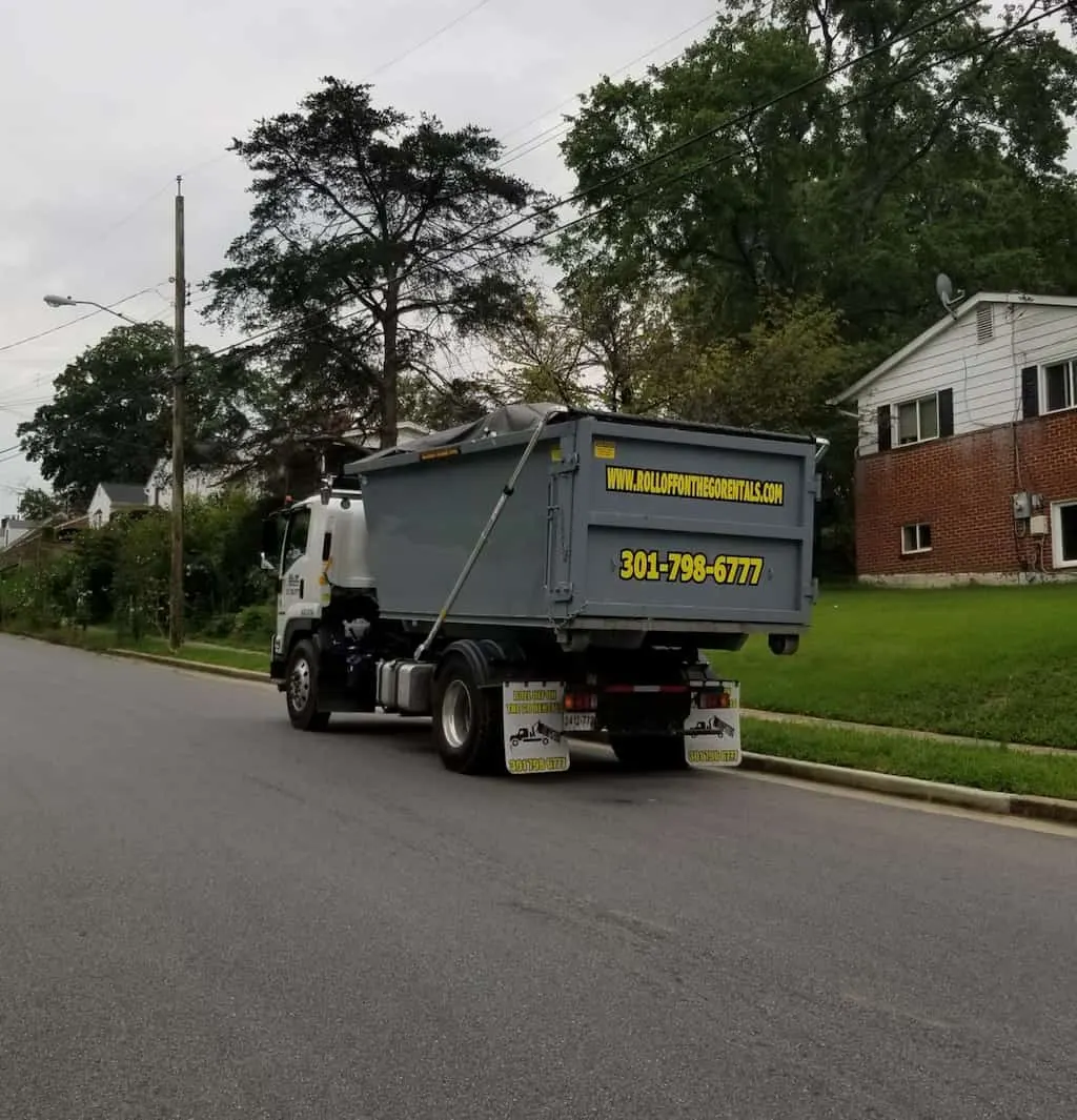 Roll Off On The Go Rentals hook-lift truck hauling a gray roll-off dumpster marked 301-798-6777 down a residential street.