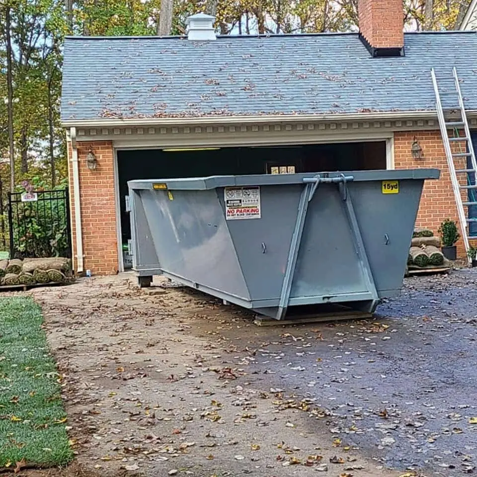 Gray 15-yard roll-off dumpster set on boards in front of an open garage at a brick home, with a ladder and sod rolls nearby.