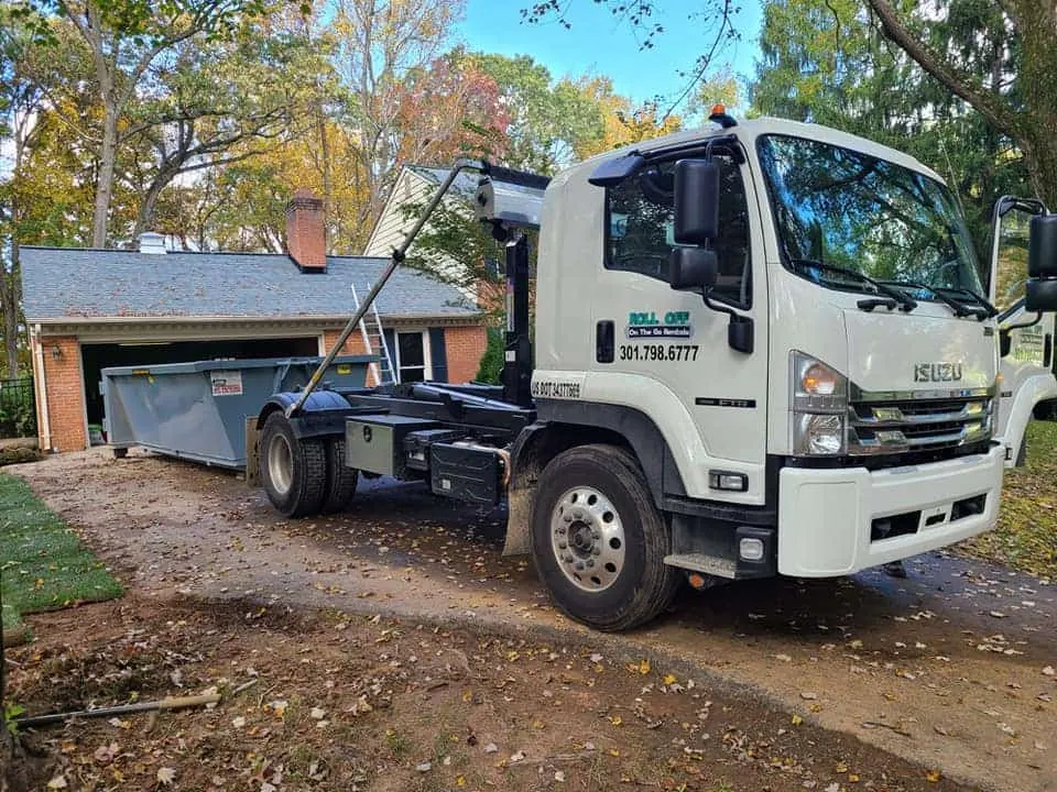 White Roll Off On The Go Rentals hook-lift truck parked in a leafy driveway next to a gray roll-off dumpster outside a brick garage.