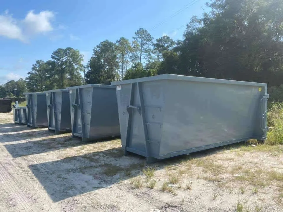 Row of empty gray roll-off dumpsters of different sizes lined up on a sandy lot in front of trees.