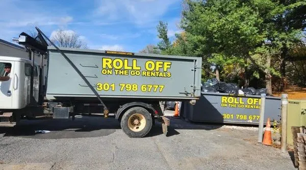 Roll Off On The Go Rentals truck carrying a gray roll-off dumpster, with a second branded dumpster full of bagged trash behind it.
