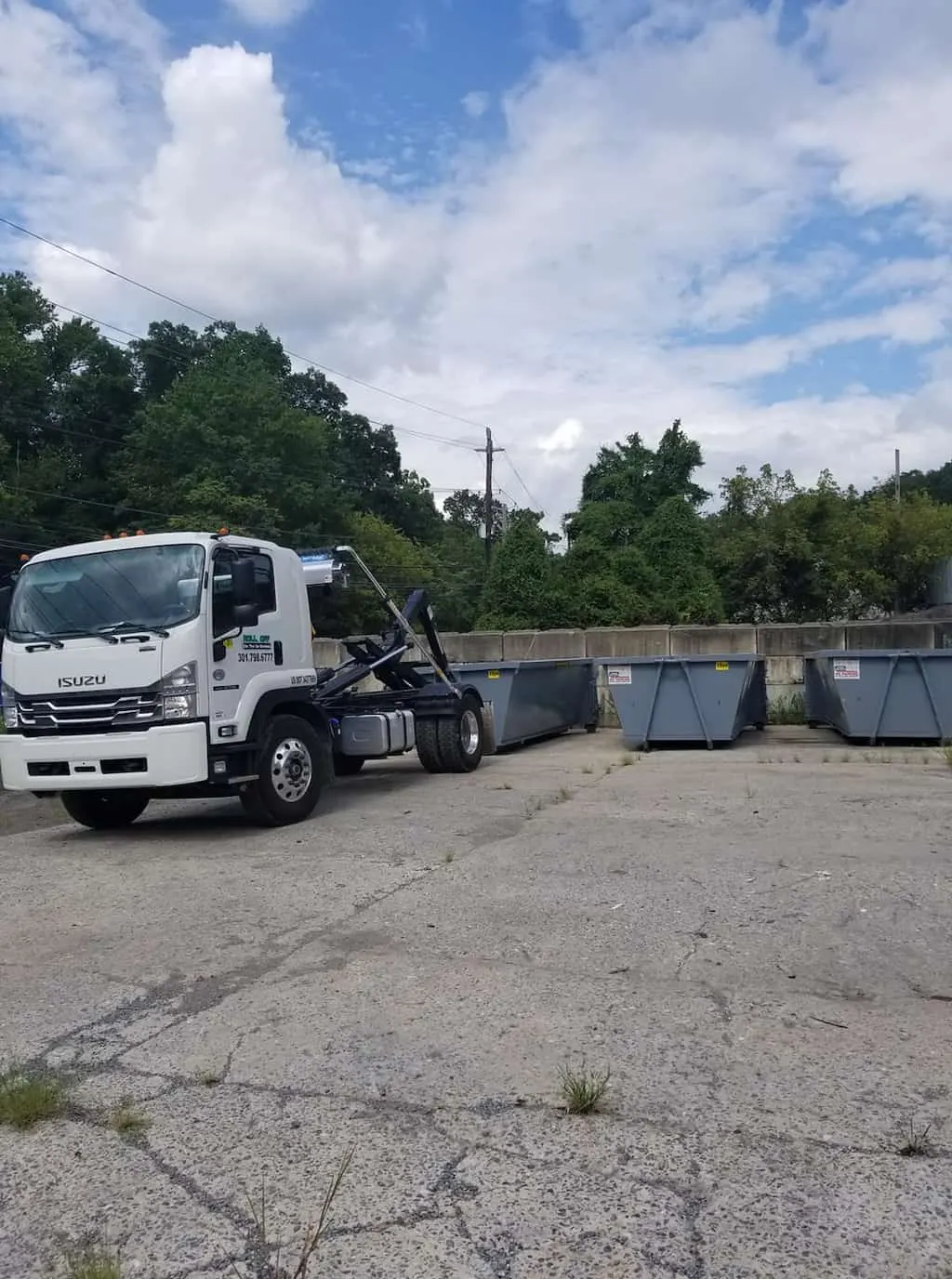 White hook-lift truck in a paved yard next to several gray roll-off dumpsters under a partly cloudy sky.