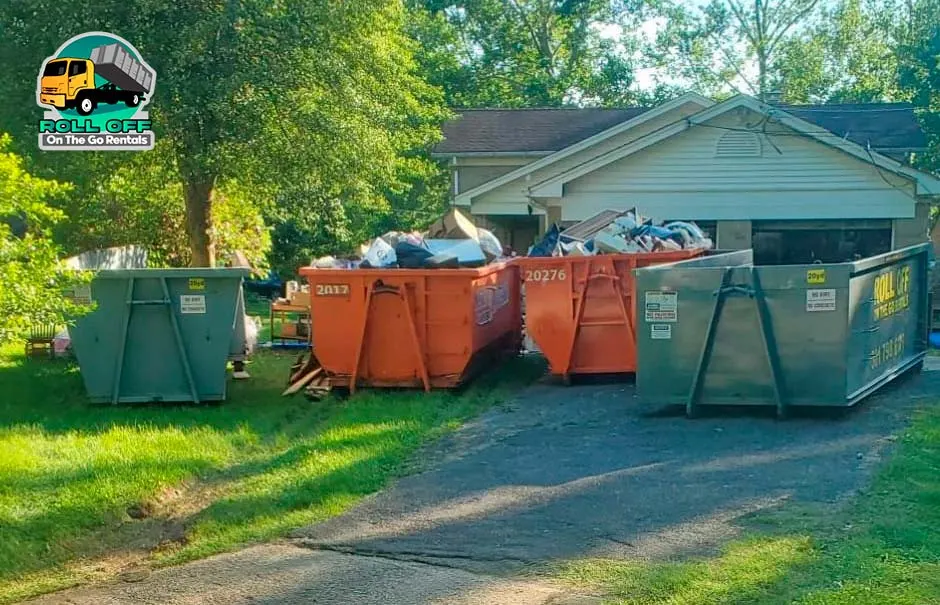 Four roll-off dumpsters, two gray and two orange and filled with household debris, lined up on a driveway in front of a house.