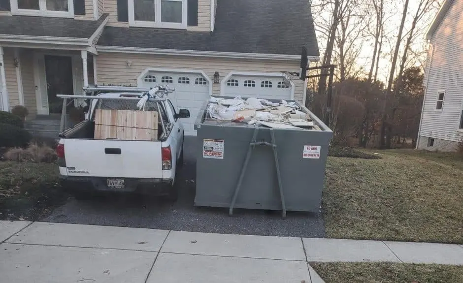 Gray roll-off dumpster loaded with construction debris in a residential driveway beside a white work pickup truck.